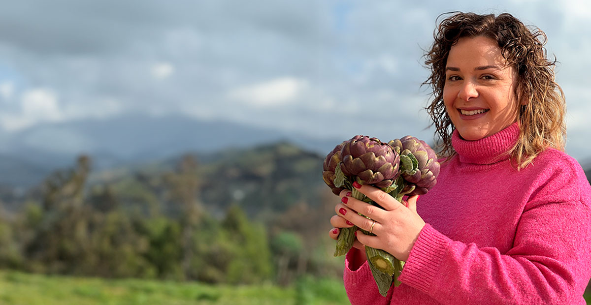 Panorama con Valentina a destra che indossa un maglione a collo alto fucsia mentre sorridente tiene in mano tre carciofi romaneschi