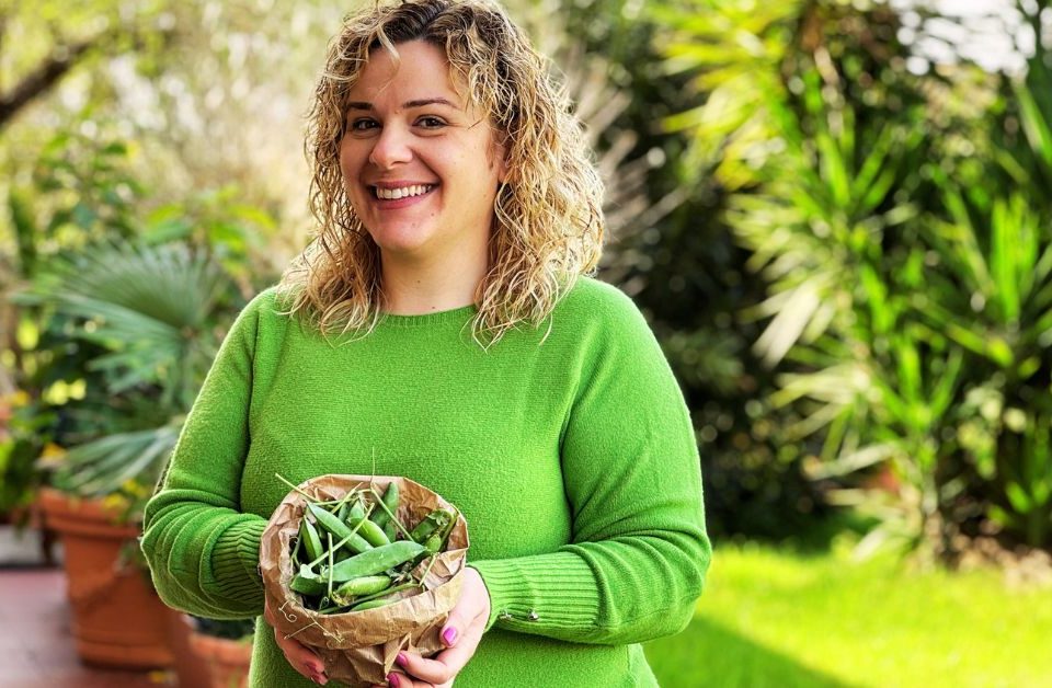 Valentina sorridente tiene in mano una piccola busta con i piselli da sgranare. Indossa un maglione verde e sullo sfondo si vedono le piante del suo giardino