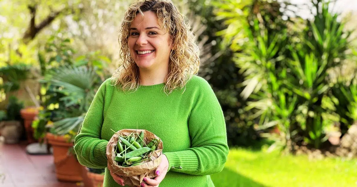 Valentina sorridente tiene in mano una piccola busta con i piselli da sgranare. Indossa un maglione verde e sullo sfondo si vedono le piante del suo giardino