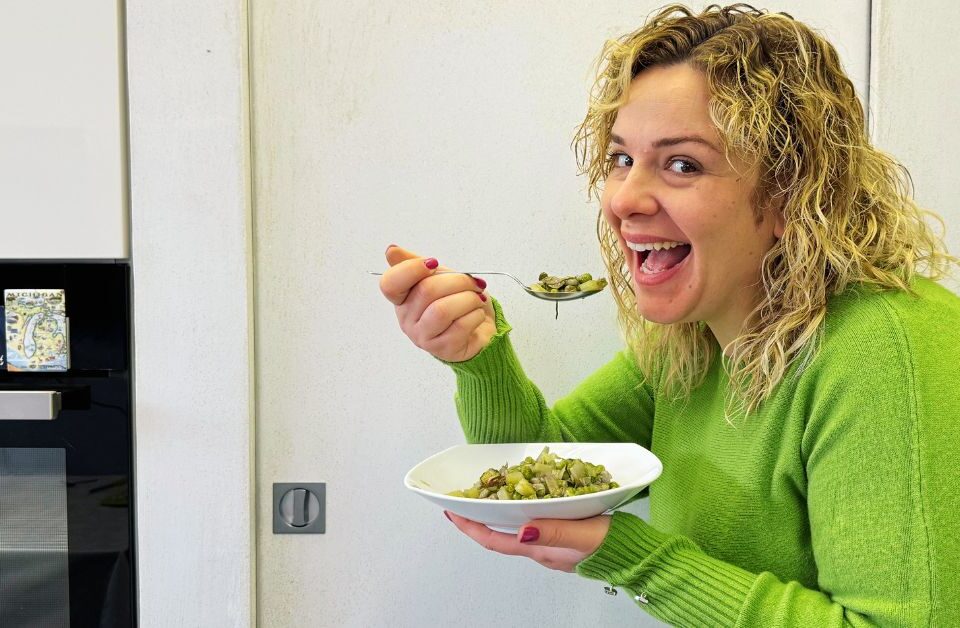 Ragazza bionda sorridente con maglia verde che sta per assaggiare la vignarola che sta nel piatto che tiene in mano.